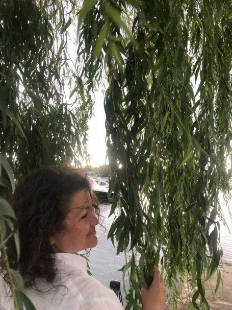 Frau steht unter einem Weidenbaum, lächelt und hält eine Zweighand.